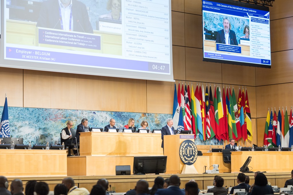 A panel of speakers at the International Labour Conference, featuring flags from various countries in the background. A speaker stands at a podium while others sit at a long table, with a screen displaying the session details.